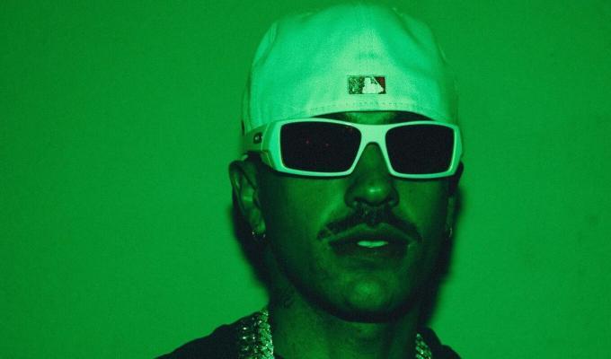 Feid con fondo verde, gorra y lentes 